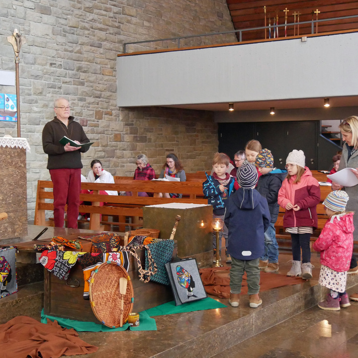 Im Altarraum der Kirche in Dipbach stehen zwei Erwachsene und zwischen ihnen mehrere Kinder. Es ist ein buntes Bild mit Stoffen und Gegenständen aus Mkoha auf dem Boden.