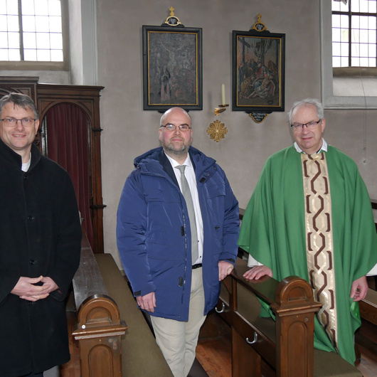 Drei Männer stehen in Kirchenbänken. Hinter ihnen an der Wand hängen zwei der Kreuzwegbilder.