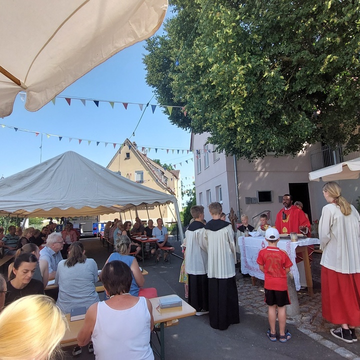 Das Bild zeigt in der Mitte den Altar, um den sich der Priester und die Ministrant:innen versammeln. In der linken Hälfte des Bildes sitzen Menschen an Biertischen.