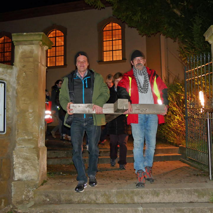Es ist dunkel. Zwei Männer tragen die Querbalken eines Holzkreuzes. Im Hintergrund sieht man die Krirche St. Vitus mit ihren erleuchteten Fenstern.