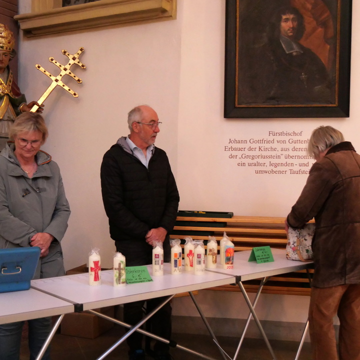 Hinter einem Verkaufstisch mit Osterkerzen stehen eine Frau und ein Mann. Davor schaut eine Frau die Kerzen an. Diese Frau sieht man nur von hinten.
