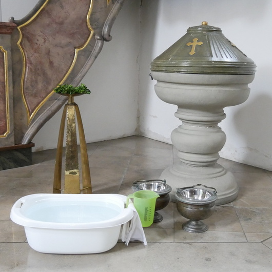 Neben dem Taufstein in der Kirche steht eine kleine Wanne mit Wasser. Daneben steht ein Schöpfbecher.